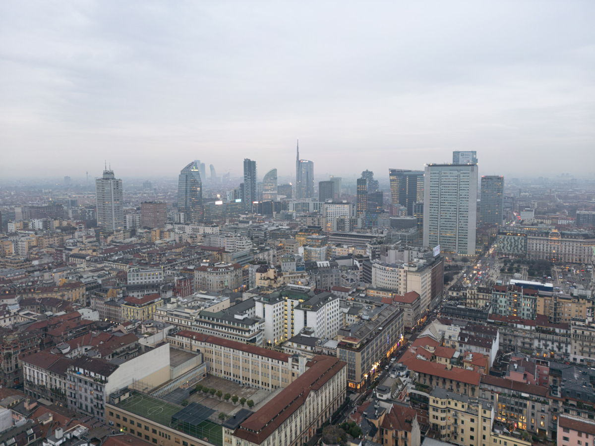 A dense layer of gray smog blankets the Milan skyline during the afternoon, obscuring the horizon behind the city's modern architectural landmarks. As Italy's financial hub transitions into the evening, the leaden sky highlights the ongoing struggle with urban air quality and stagnant weather patterns.