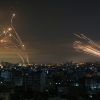 Against a dark night sky, Israel's Iron Dome missile defense system, appearing as lights high in the sky on the left, intercepts rockets shooting through the sky from the right. On the ground are multistory buildings with illuminated windows.