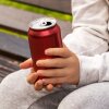 In this photo, a child holds a drink in a dark red aluminum can. This child is seated and is wearing gray pants and a long-sleeved gray shirt.