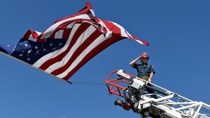A captain with the Clifton Fire Protection District salutes the passing procession carrying the bodies of three firefighters killed while fighting the Snyder Fire in Grand Junction, Colo., on June 28. He is at the top of a ladder on a fire truck and an American flag waves above him.
