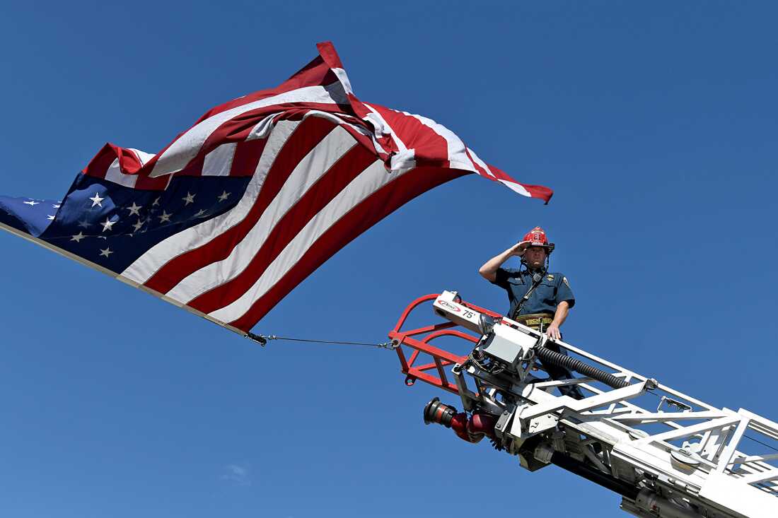 A captain with the Clifton Fire Protection District salutes the passing procession carrying the bodies of three firefighters killed while fighting the Snyder Fire in Grand Junction, Colo., on June 28. He is at the top of a ladder on a fire truck and an American flag waves above him.
