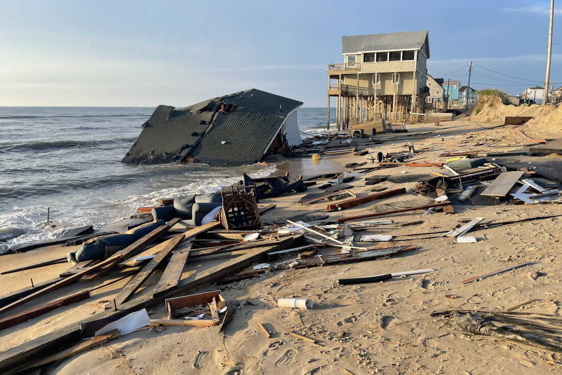 A collapsed house lies in shallow ocean water on the coast. Debris such as planks of wood and a sofa lie scattered on the sand. In the background is a two-story house that sits on top of tall wooden stilts.