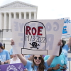 One protestor holds a handmade sign that shows a tombstone. It reads, "Roe 1973-2022" above a skull-and-crossbones drawing. The Supreme Court building is visible in the background. Another holds a sign that says, "The Future is Anti-Abortion."