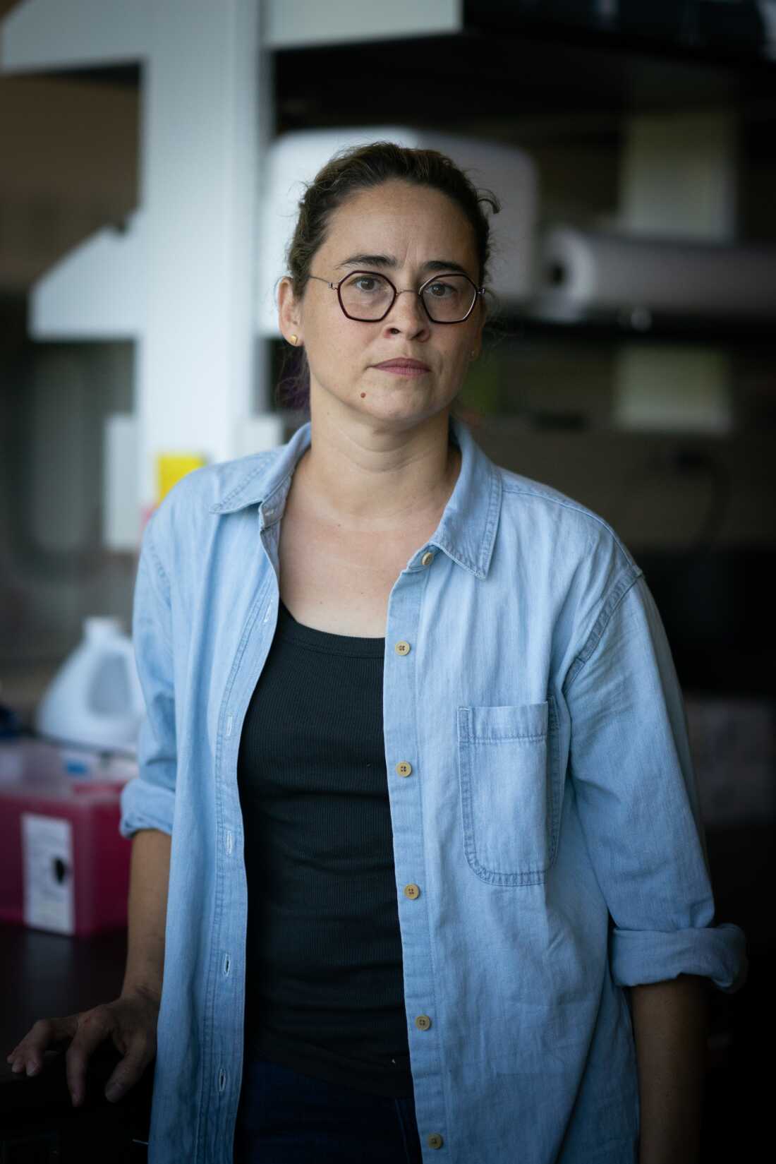 This photo shows Elisabeth Forrestel, an assistant professor of viticulture and enology at the University of California, Davis. She's wearing a black shirt with an unbuttoned light blue shirt layered on top of it. She's also wearing glasses and has her dark hair tied back. Laboratory equipment is in the background.