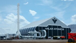 A large warehouse-like industrial building has the capitalized letter "VALAR ATOMICS" across the front. It is black with a white, pitched roof and is sitting under a blue sky with light, white clouds. Large ventilation pipes are coming from the building's front. A couple pieces of medium sized red machinery sit out front. Behind the building to the left is a mountain.