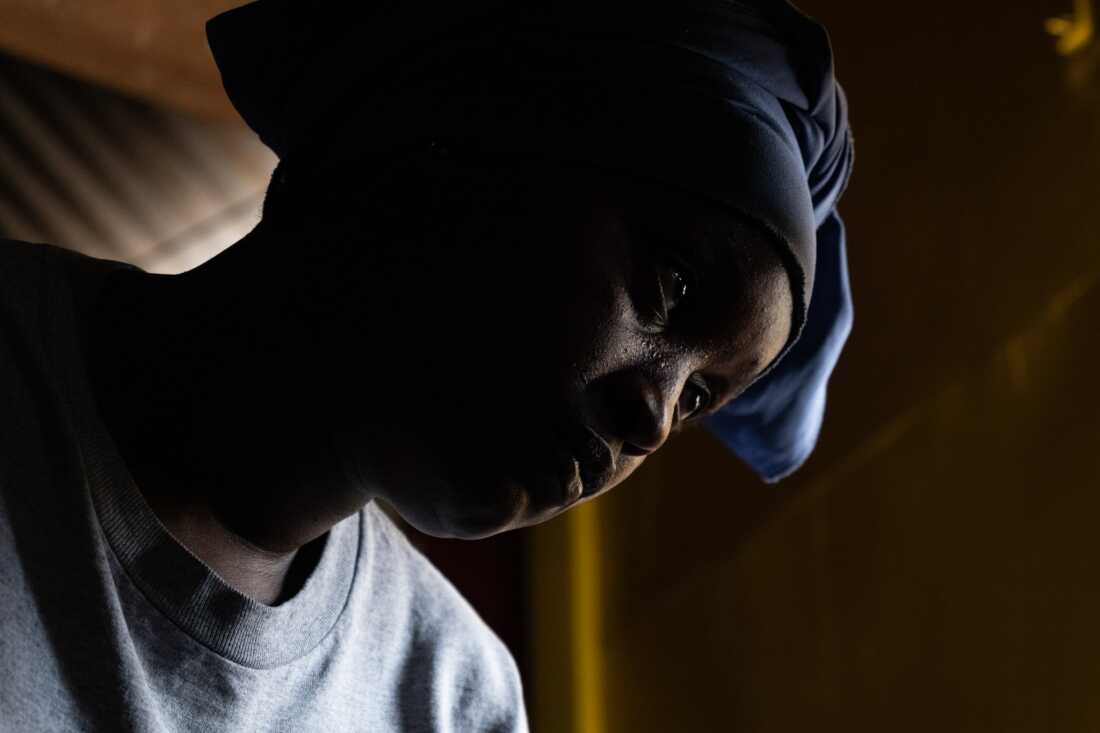 A close-up shows a woman's face and shoulders. She's wearing a gray T-shirt and a blue head covering.