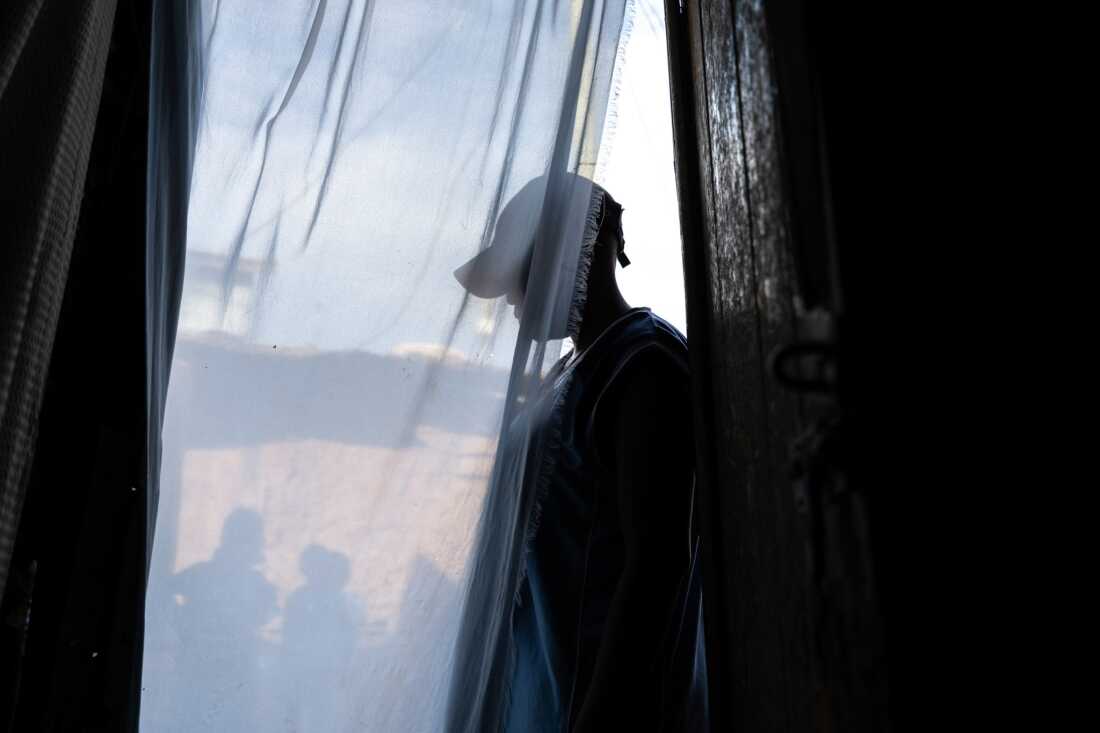 A transgender man living wearing a baseball cap is shown in silhouette in a doorway behind a sheer curtain.