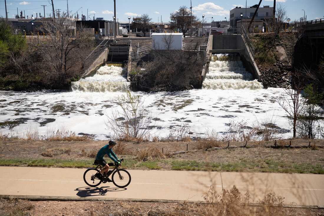 A cyclist rides a bike on a path that passes by foamy-white treated wastewater outfalls as they flow into the South Platte River. Metro Water Recovery’s treatment facility in Denver is in the background.
