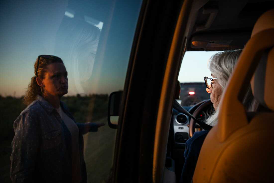 Robyn Smith sits behind the steering wheel of a vehicle as she speaks with Heather Marsh, who is standing outside the vehicle, on the left side of the frame.