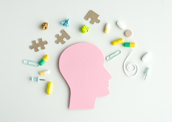 A pink head with a puzzle piece on it. There are many pills scattered around the head. Some of the pills are yellow and some are green