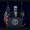 In this screenshot from a February 28 video, President Trump stands behind a lectern to discuss strikes on Iran. Speaking into a microphone, he's wearing a white baseball cap that says "USA" in navy blue capital letters on it. The presidential seal is on the front of the lectern.