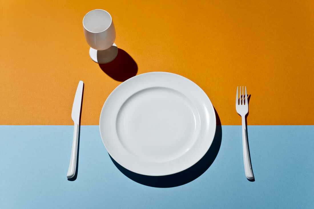 This image shows a high-angle view of a white plate, a stemmed drinking glass, a fork to the plate's right and a knife to the plate's left. The items are all set on a tabletop that's orange across the top of the frame and light blue across the bottom.