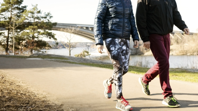 Two people wearing sneakers and photographed from the neck down walk on a paved path that runs along a body of water. A bridge is in the background.