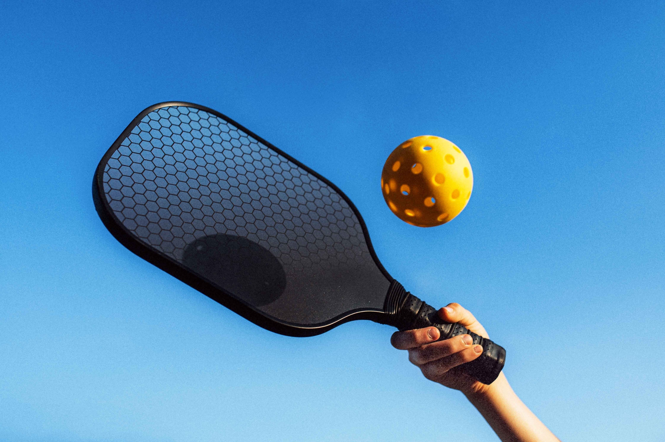 A hand holds a paddle against a clear blue sky, preparing to hit a yellow pickleball. The scene suggests a bright and sunny day, with the pickleball clearly in motion.