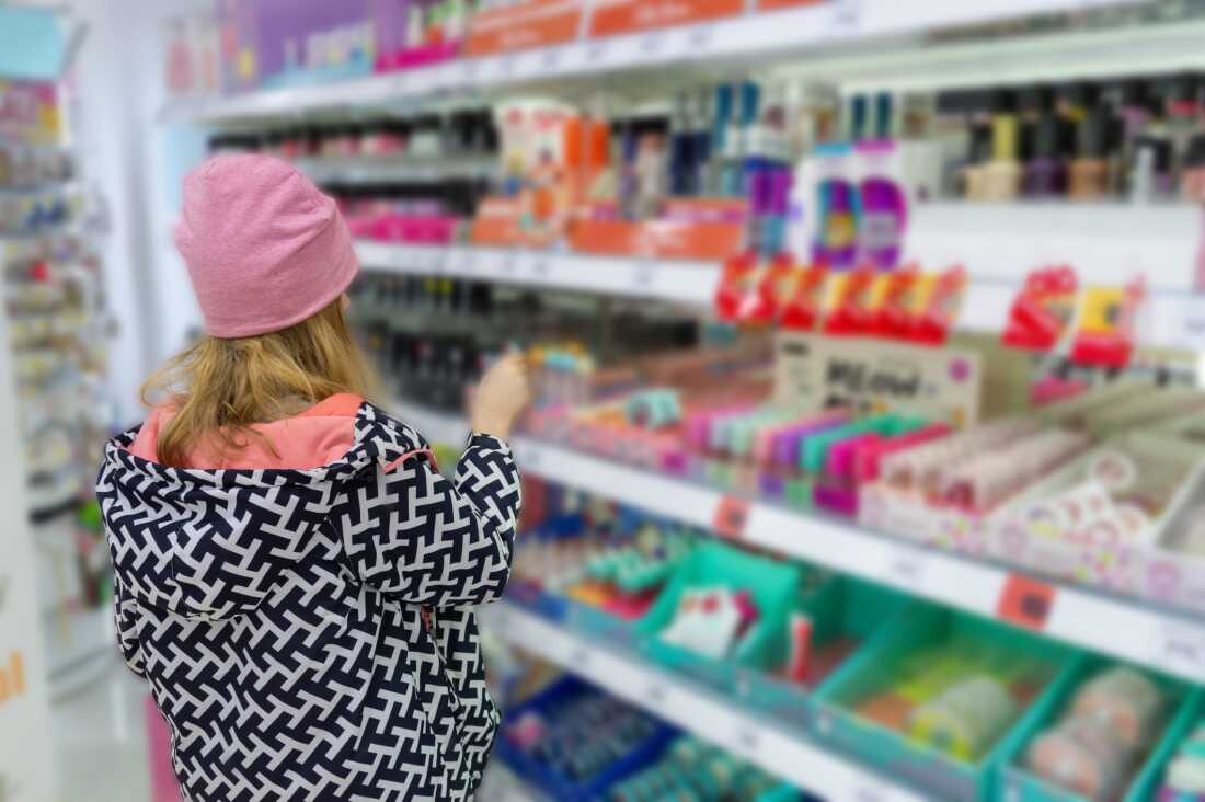 A girl with her back to the camera stands in a store, facing shelves of health and beauty products on display for sale. The girl appears to be the size of a tween or young teen and is wearing a black and white patterned coat and a pink winter cap.