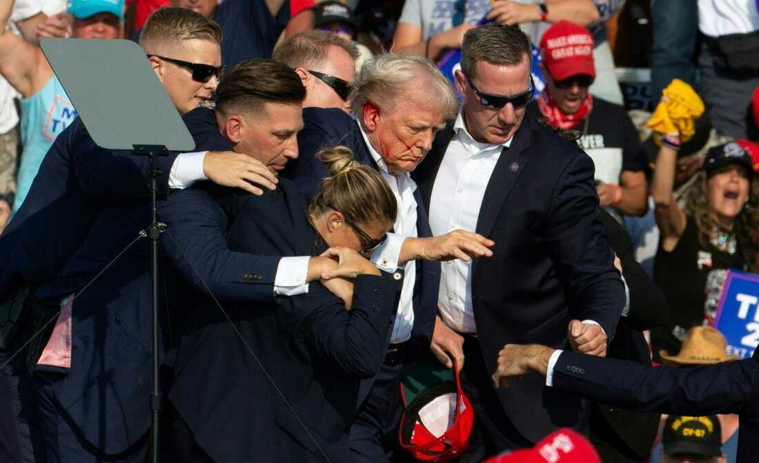 Donald Trump has blood on his face and right ear as Secret Service agents in dark suits surround him and help him off the stage amid the assassination attempt.