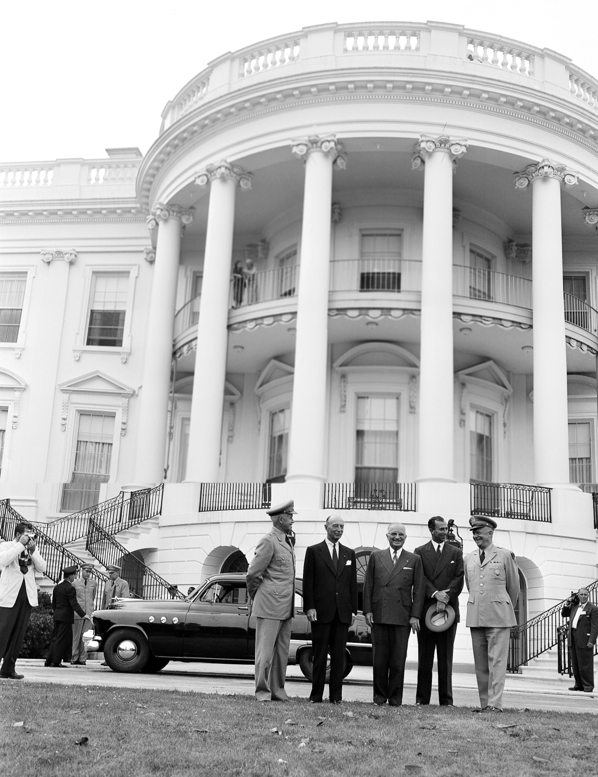 As President Harry Truman and General Dwight Eisenhower greet military officials at the White House in June 1952, First Lady Bess Truman and First Daughter Margaret Truman hang over the railing on the upper balcony.