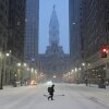 A person wearing a coat and boots walks across a snowy street during a winter storm in Philadelphia on January 25. Tall buildings rise on both sides of the photo.