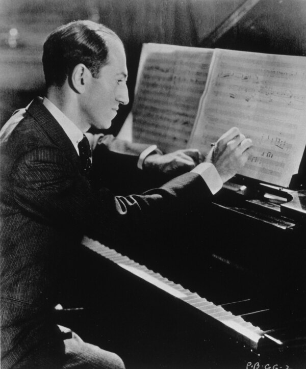 George Gershwin writes sheet music while sitting at a piano.
