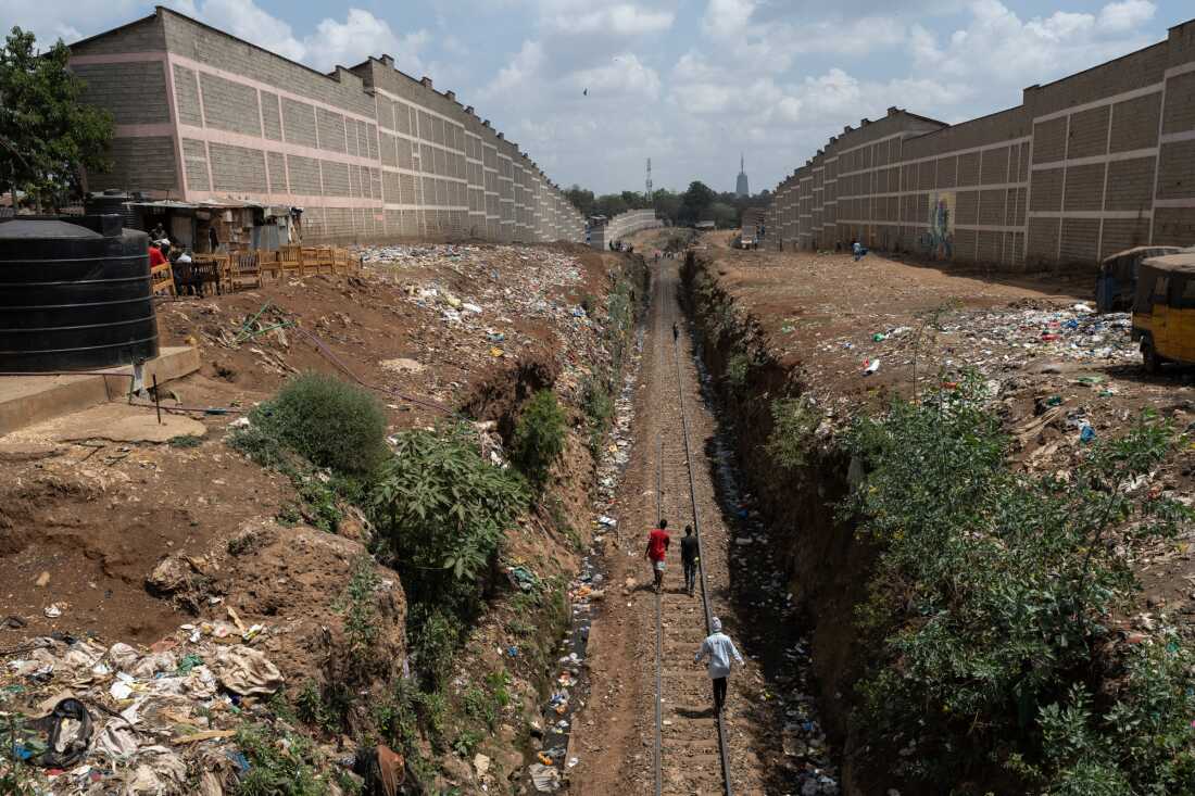 People walk along train tracks running between two high walls. There's trash on the ground all around.