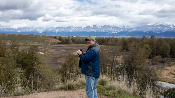 Mike Durglo Jr. holds binoculars and looks toward the camera. He stands before a mountain range in Montana. 