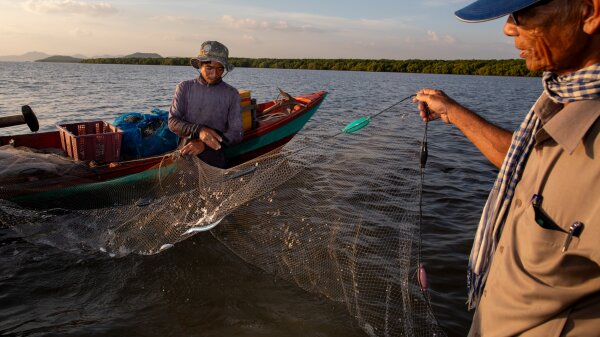 A man on a green boat holds one end of a fishing net above the water and another man holds onto the other side of the net.