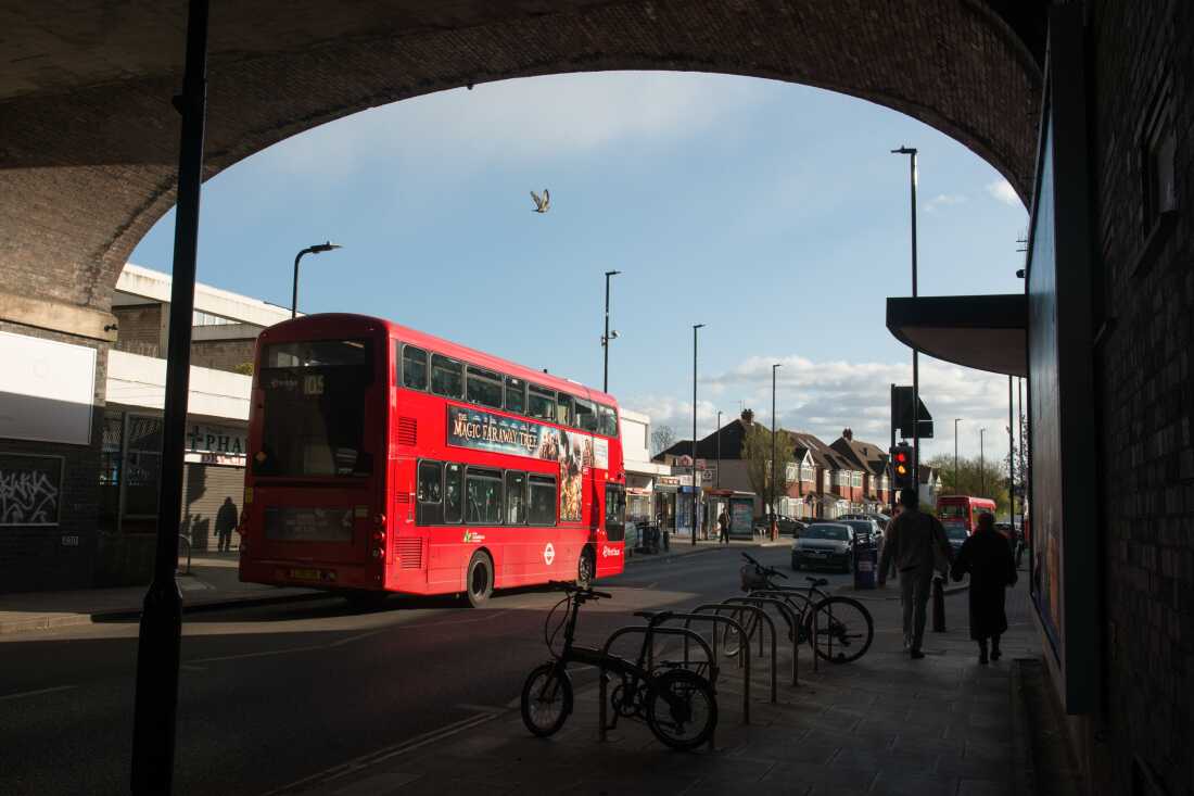 A red double-decker bus passes underneath a train bridge arch at Greenford Tube station.
