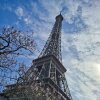 A low-angle view of the Eiffel Tower, partially framed by flowering branches under a blue sky dotted with clouds in Paris, France, on April 10, 2026.
