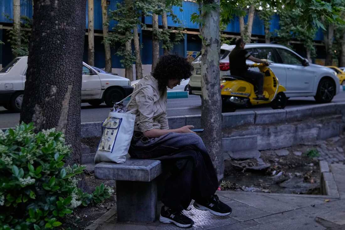 A woman checks her smartphone while sitting on a bench along a sidewalk in northern Tehran, Iran, Tuesday, May 26, 2026.