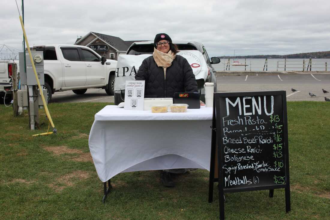Chloe Chalakani is shown in a wide-angle photo standing at her table at a farmers market where she sells specialty pasta. The board shows her offers and prices. 