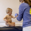 A child, who appears to be about a year old, is sitting on an exam table, wearing a diaper. A medical professional is holding the child by the hand.