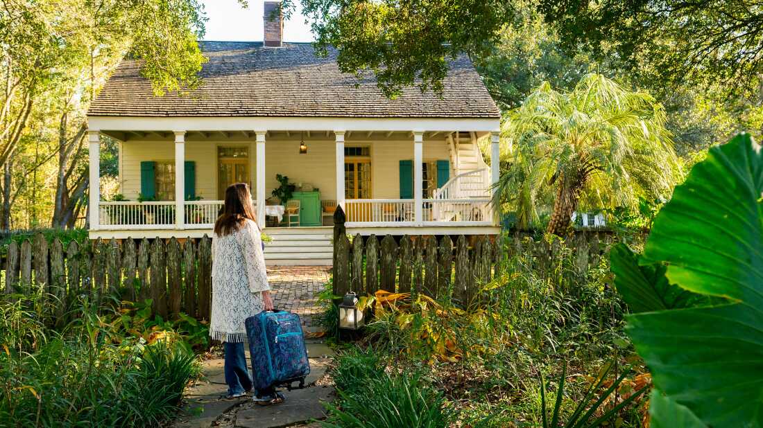 A rearview photograph of a person holding a rolling suitcase as they stand on a stone pathway facing a yellow cottage with a quaint front porch. The person walks along the stone path, which is framed by lush foliage, approaching the house where they will stay as a houseguest.