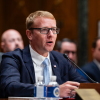 A man in a dark suit, white shirt and blue tie, with short hair and wearing glasses, sits at a table in a congressional hearing room and speaks into a microphone.