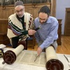 A man reads from the Torah using a traditional pointer to follow the words, while the rabbi looks on, holding a microphone for the reader.