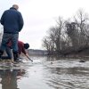 Two men take water samples from the Potomac River in Maryland in January. One man is wearing blue disposable gloves and is crouching down to reach into the river to collect a sample.