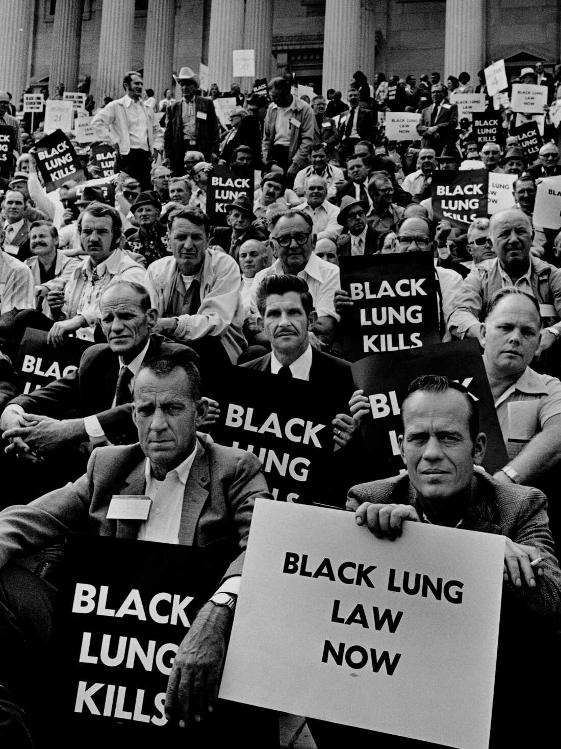 In this 1975 black-and-white photo, coal miner men sit and stand on the U.S. Capitol steps holding signs with phrases such as "Black Lung Kills."