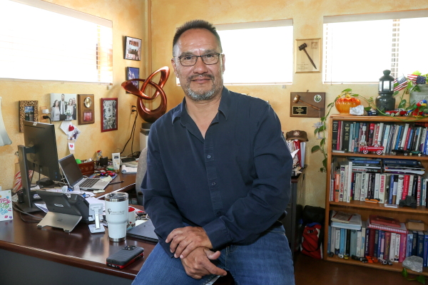 Fernando Shipley perches on a desk in an office packed with books, plants and pictures on the walls. He is smiling and wearing glasses.