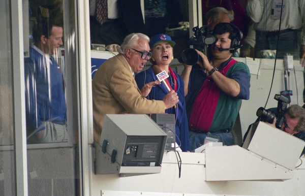 First lady Hillary Rodham Clinton, right, and Chicago Cubs announcer Harry Caray, left, sing "Take Me Out To The Ball Game" during the seventh inning stretch at Wrigley Field in Chicago in 1994. The Cubs are leaving WGN Radio that's been their radio home for 90 years. Radio president Jimmy de Castro confirmed on the air Wednesday, June 4, 2014, media reports that the Cubs are leaving the station after this season. (AP Photo/John Zich, File)