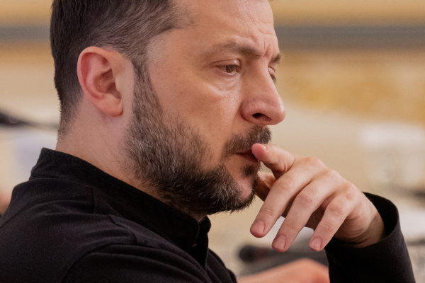 This photo shows a side view of Ukrainian President Volodymyr Zelenskyy from the shoulders up. He's wearing a black shirt, and one finger is touching his upper lip.