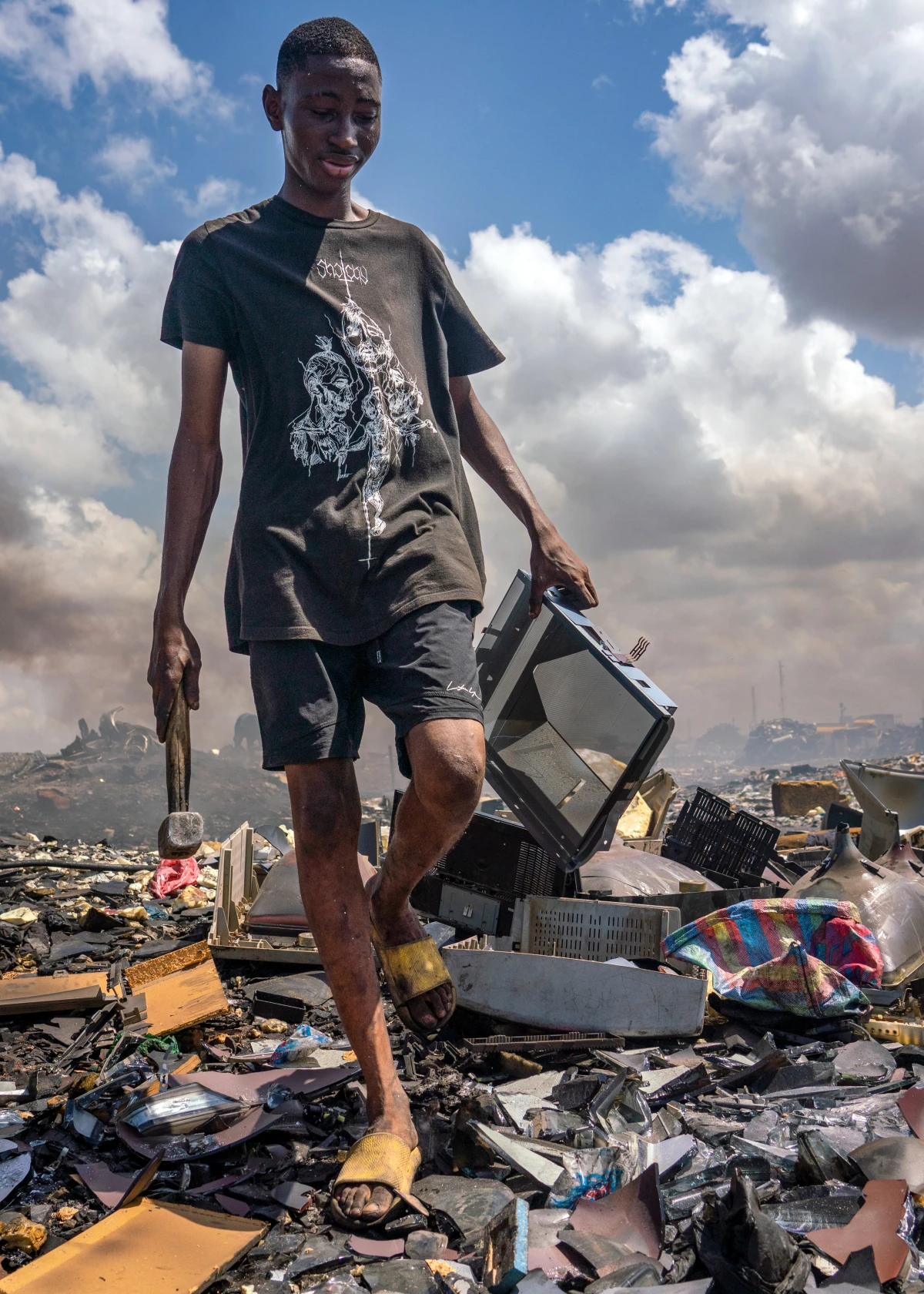 An 18-year-old holding a hammer and a TV walks over pile of e-waste. Collecting e-waste to salvage can be dangerous work. Cuts, burns and other injuries are common, and e-waste sites are often blanketed in noxious fumes.