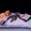A pink mug of hot tea, a gray silk sleep eye mask, two books and some wired headphones are laid out on top of two stacked pillows next to a table lamp that's turned on, emitting warm light. All the objects are photographed in a studio against a black backdrop.