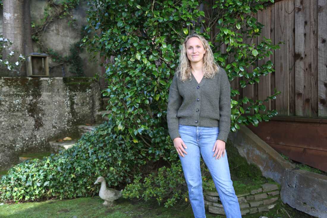 Marta Galic stands outdoors on grass in an area with green foliage, a concrete wall and a wooden fence in the background. She is wearing blue jeans and a dark gray, button-down sweater.