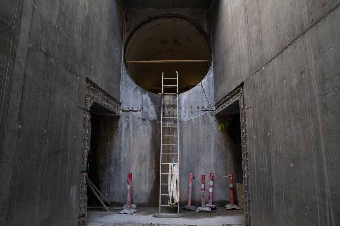 This photo shows a large-diameter tunnel — the Kalvebod Brygge tunnel project — under construction. A tall ladder extends upward to the bottom of the tunnel's circular opening. The bottom of the ladder is in an area with a concrete floor and tall concrete walls on the left and right.