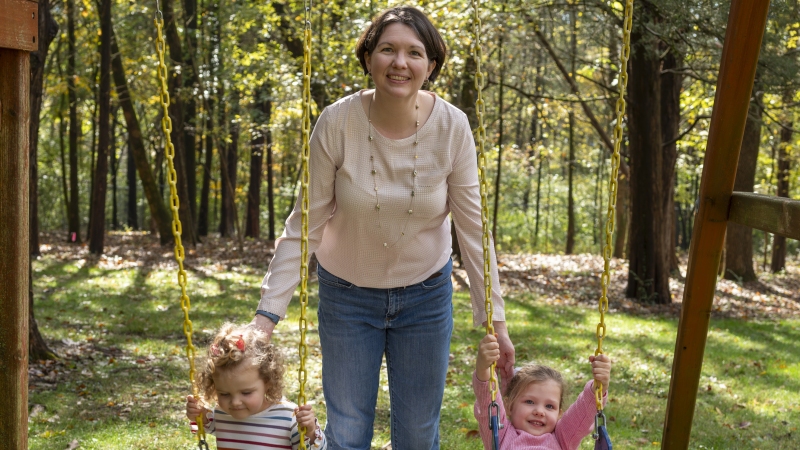 Laura Terry stands in between her two daughters, who are seated on a swing set.