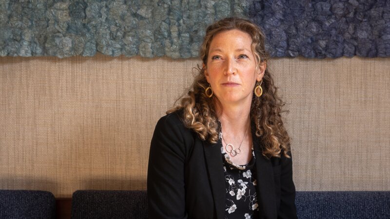 Noella Sudbury is the founder of Rasa Legal, a non-profit that uses technology to help people expunge their criminal records. She is sitting on a couch with her hands in her lap. Sudbury has long, curly hair and she is wearing a dark suit and a dark floral top. 