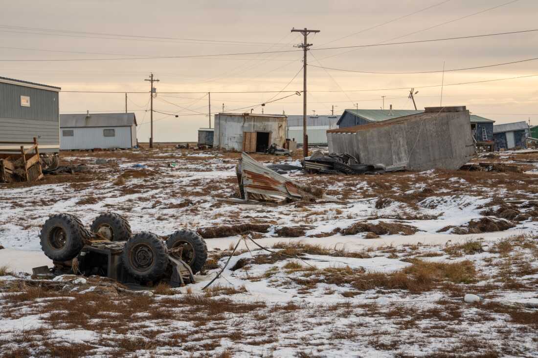 A shed-like building and an all-terrain vehicle lie overturned upside down in Kwigillingok, Alaska, in late October. The ground is partially covered by snow. In the background stand a few small, one-story buildings. Power lines supported by poles crisscross the area.