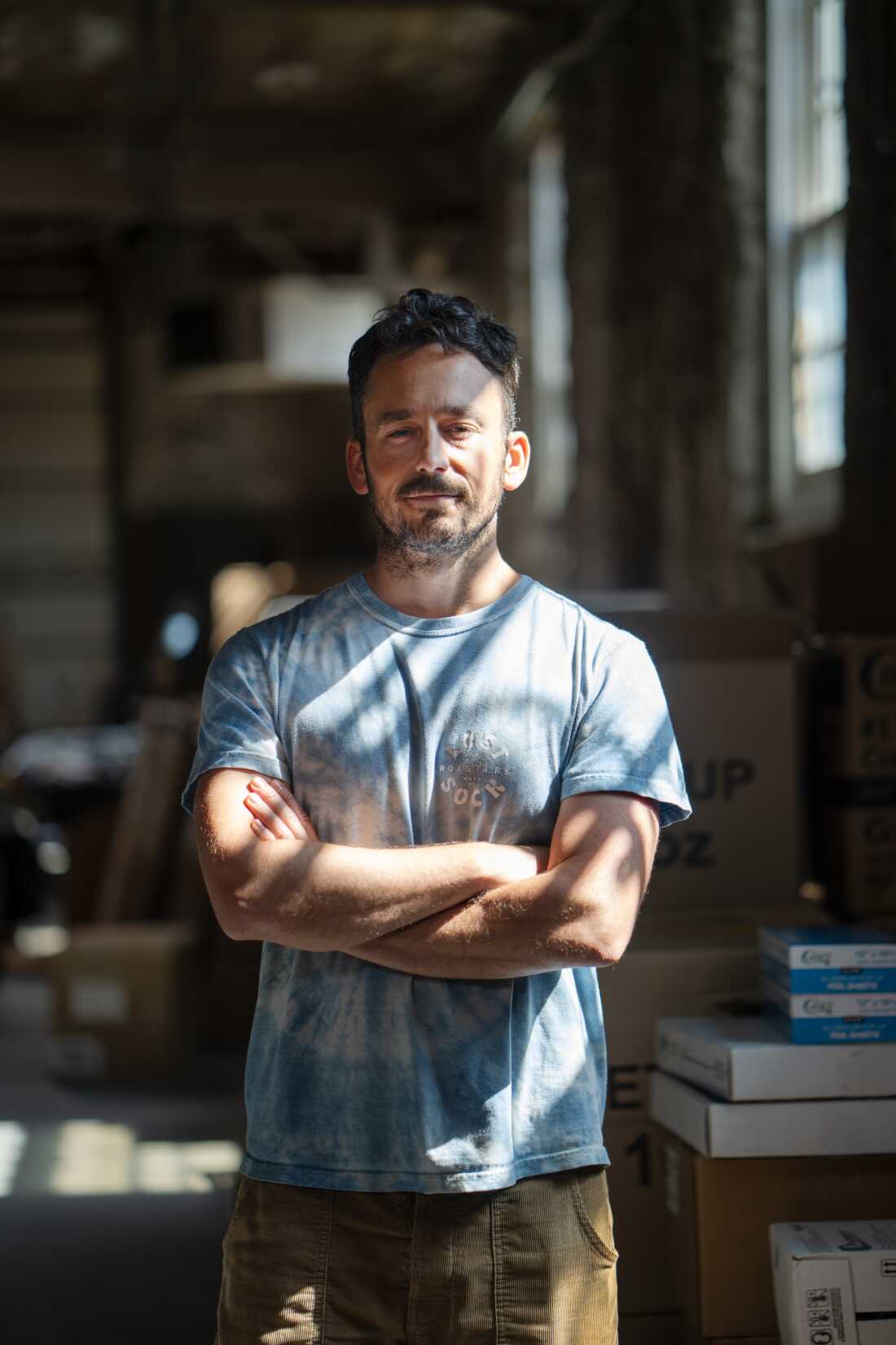This photo shows Jeff Yerxa at Lost Sock Roasters in Washington, D.C. He's standing with his arms crossed over his torso, and he's wearing a light blue T-shirt and camel-colored corduroy pants.