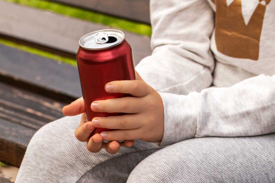 In this photo, a child holds a drink in a dark red aluminum can. This child is seated and is wearing gray pants and a long-sleeved gray shirt.