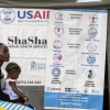 In this photo, 20-year-old Chiedza Emmanuel is seated at a clinic in Zimbabwe in 2024 as she waits to undergo a contraceptive implant procedure. A young girl is sitting in her lap. A large banner displays different contraceptive methods, and another banner bears the logo of the U.S. Agency for International Development.