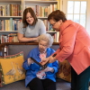 Caroline Brown (left) and Cynthia Briscoe Brown look at a family photo album with their grandmother and mother Barbara Briscoe (seated) in Atlanta. Caroline, 33, wears a gray t-shirt and stands behind a vintage sofa. Cynthia, 65, wears a pink blazer and holds a photo album while Barbara, 93, sits. Barbara wears a blue shirt over a pink patterned shirt.